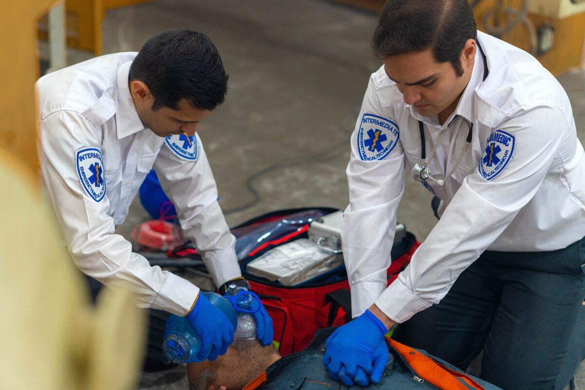 Two first responders and EMS staff in white uniform shirts and blue gloves perform CPR on a Electrocution victim. One is providing CPR, the other is using a bag valve mask.