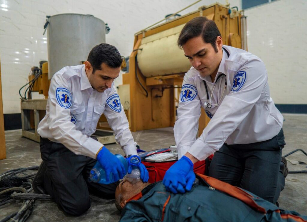 Two first responders and EMS staff in white uniform shirts and blue gloves perform CPR on a Electrocution victim. One is providing CPR, the other is using a bag valve mask.